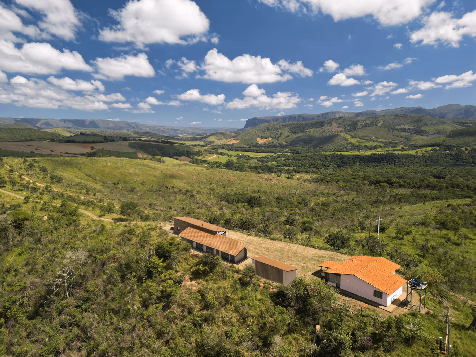 Aerial view of rural houses with terracotta roofs surrounded by green hills and forests under a partly cloudy blue sky.
